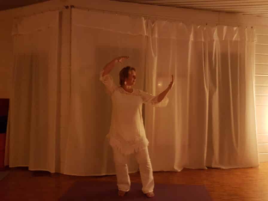 Heike Engel in weißer Kleidung in einer Qigong-Haltung mit erhobenen Armen in einem hellen Raum