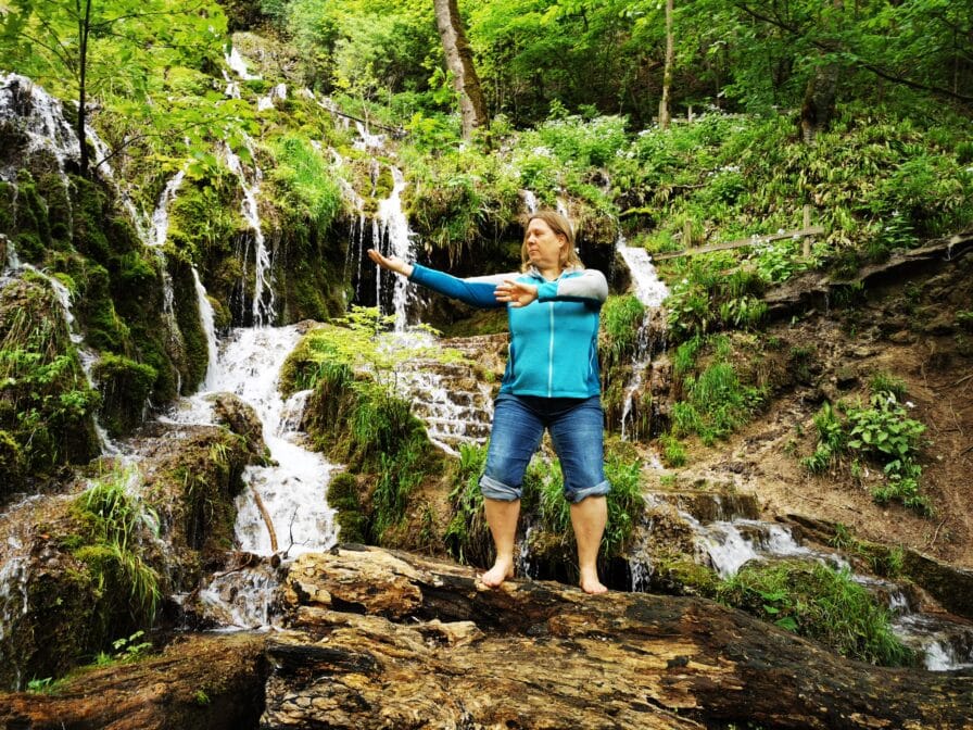 Heike Engel übt Qigong mit ausgebreiteten Armen auf einem Felsen vor einem Wasserfall im Wald