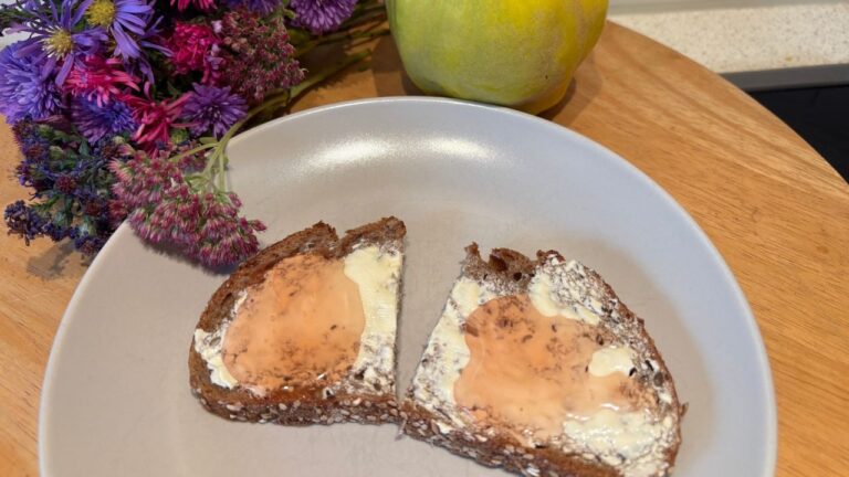 Zwei Scheiben dunkles Brot mit Butter und Quittengelee auf einem Teller, daneben Herbstblumen und eine Quitte