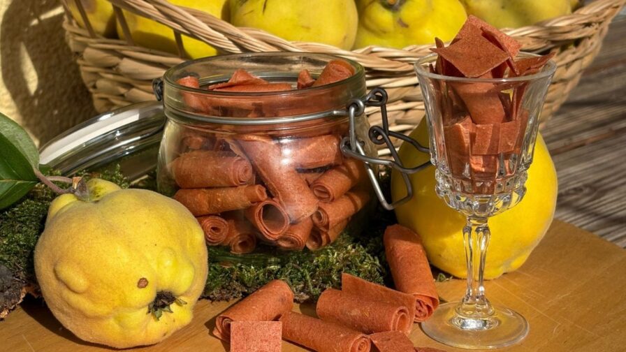 Aufgerollte Stücke Quittenleder in einem Glas und einem Stielglas, daneben frische Quitten und ein Korb