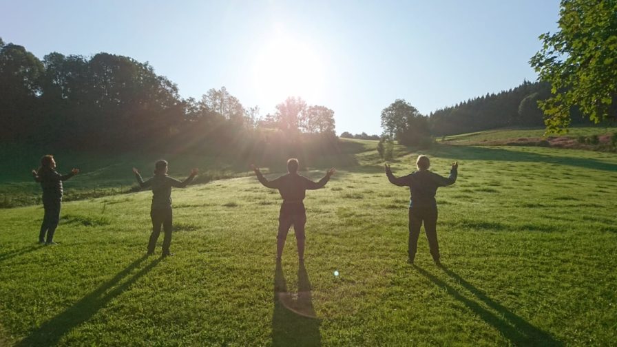Drei Menschen üben Qigong mit ausgebreiteten Armen im Gegenlicht der Morgensonne auf einer Wiese